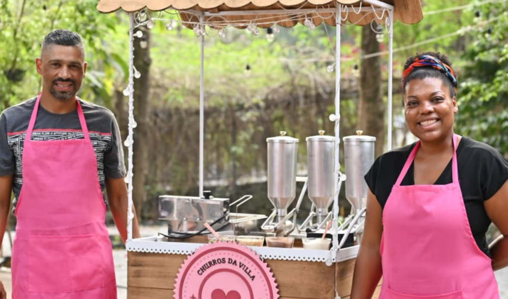 Imagem do carrinho em tom bege e rosa do Churros da Villa. Do lado esquerdo o Fabiano e a Suellen, ambos usam camiseta e um avental rosa. Ao fundo muro com árvores.