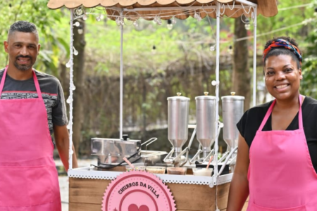 Imagem do carrinho em tom bege e rosa do Churros da Villa. Do lado esquerdo o Fabiano e a Suellen, ambos usam camiseta e um avental rosa. Ao fundo muro com árvores.