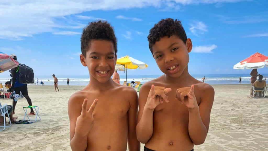 Nicolas e Miguel são duas crianças de pele negra, estão aparecendo na foto da barriga para cima na praia. Ao fundo céu azul com diversas nuvens. Três guarda-sol coloridos e pessoas na imagem. 