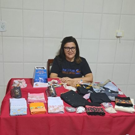 Mulher da AMAMOS está sentada numa mesa expondo trabalhos manuais e outros produtos numa mesa de toalha vermelha.