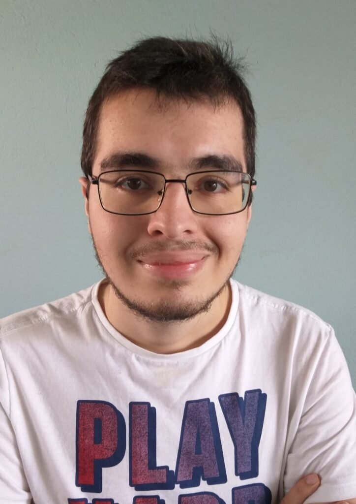 Foto perfil do Luciano, homem branco, cabelos e olhos castanhos, usa óculos de grau. Tem barba rala e usa camiseta branca com uma estampa com a palavra play.