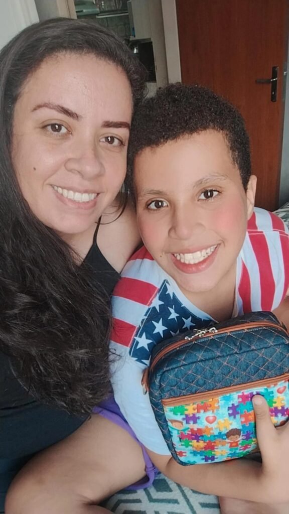 Selfie da Raquel e seu filho Vitor e ele segura uma bolsa pequena em jeans e tecido com quebra-cabeças do autismo. 