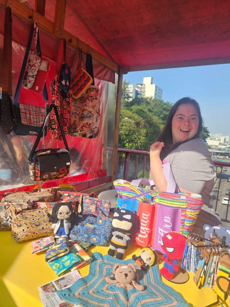 Bia, mulher de pele branca, cabelos castanhos cumpridos, está de lado com uma bolsa em seu braço. Ela está sentada em um stand montado a céu aberto. Na mesa diversos produtos da sua marca. Na lateral Uma parede plástica com diversas bolsas penduradas. Ao fundo, diversos carros estão na avenida passando embaixo da ponte onde estão expondo. 