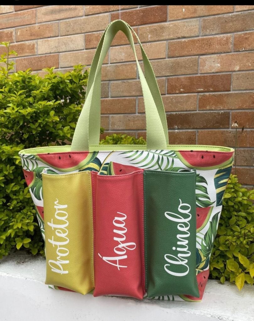 Bolsa grande de tecido com fundo floral e de melancia com local externo para colocar o protetor, água e o chinelo, pois possuem texto em letra branca na lateral. 